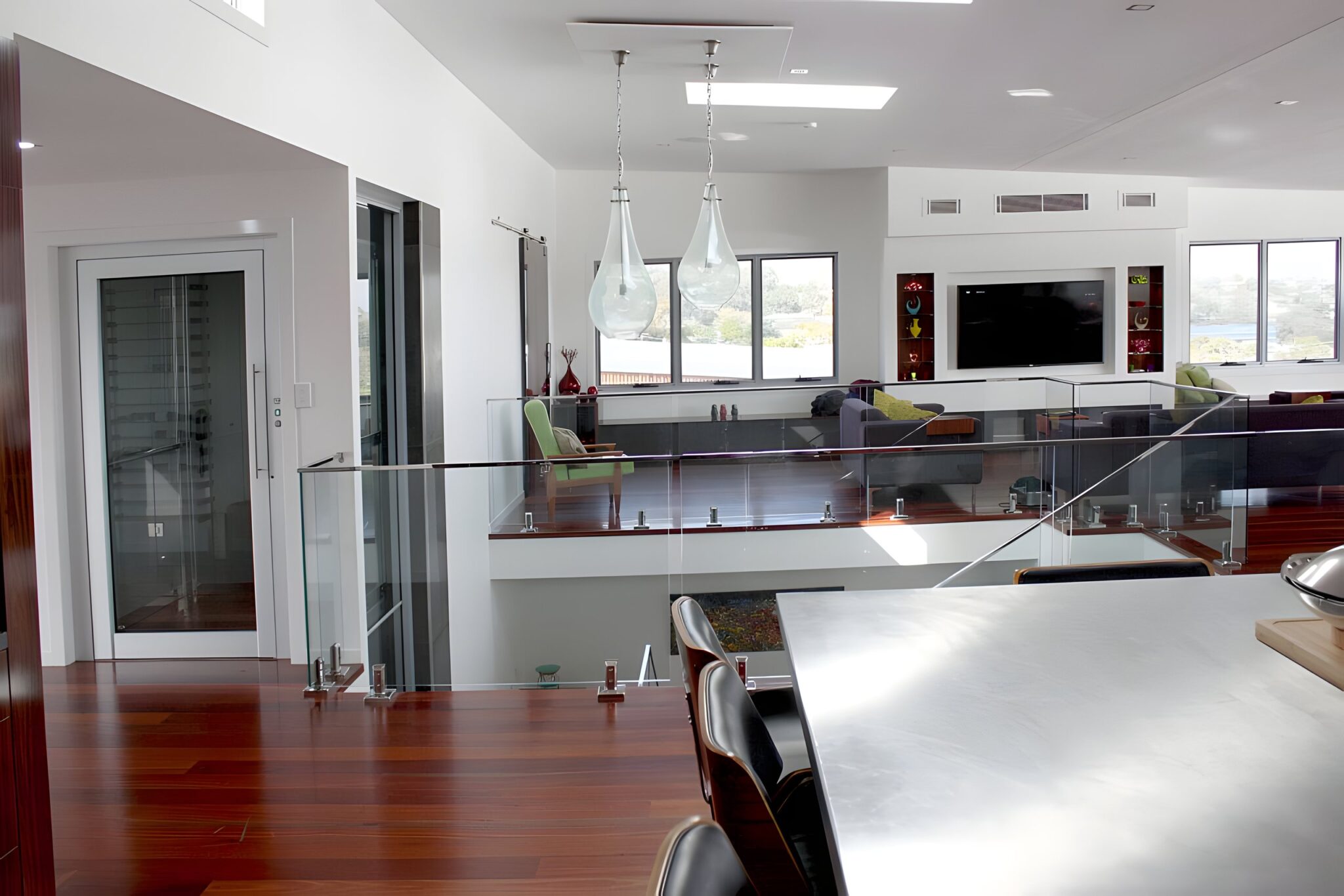 Frosted glass lift door beside a glass balustrade overlooking the open-plan living area of a hillside home in Morningside, Brisbane