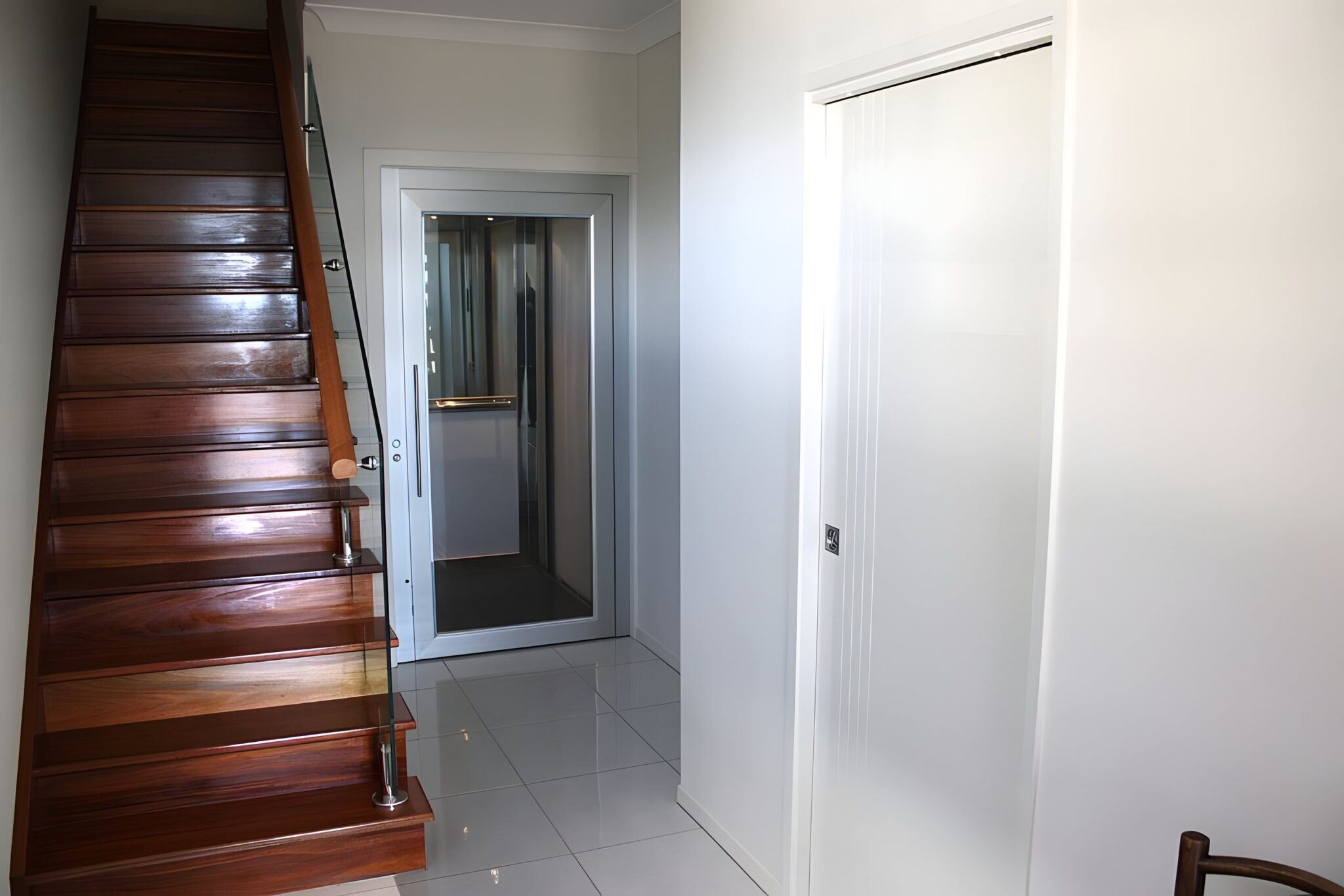 Timber staircase with a glass balustrade beside a white hallway with a glass-panelled lift door at the far end, Brisbane