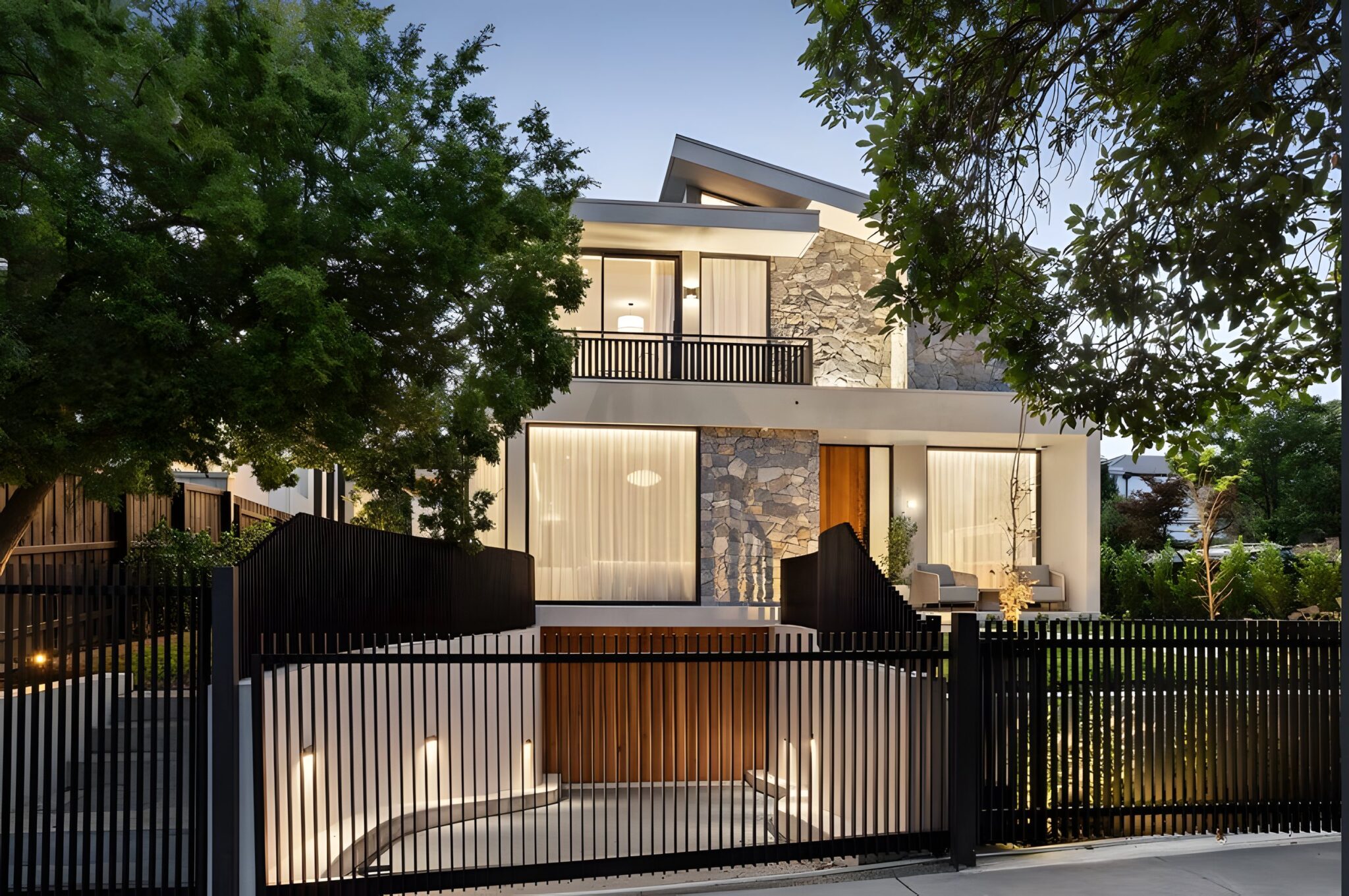 Architect-designed three-level home in Kew at dusk with a stone feature wall, timber screening and a black fence