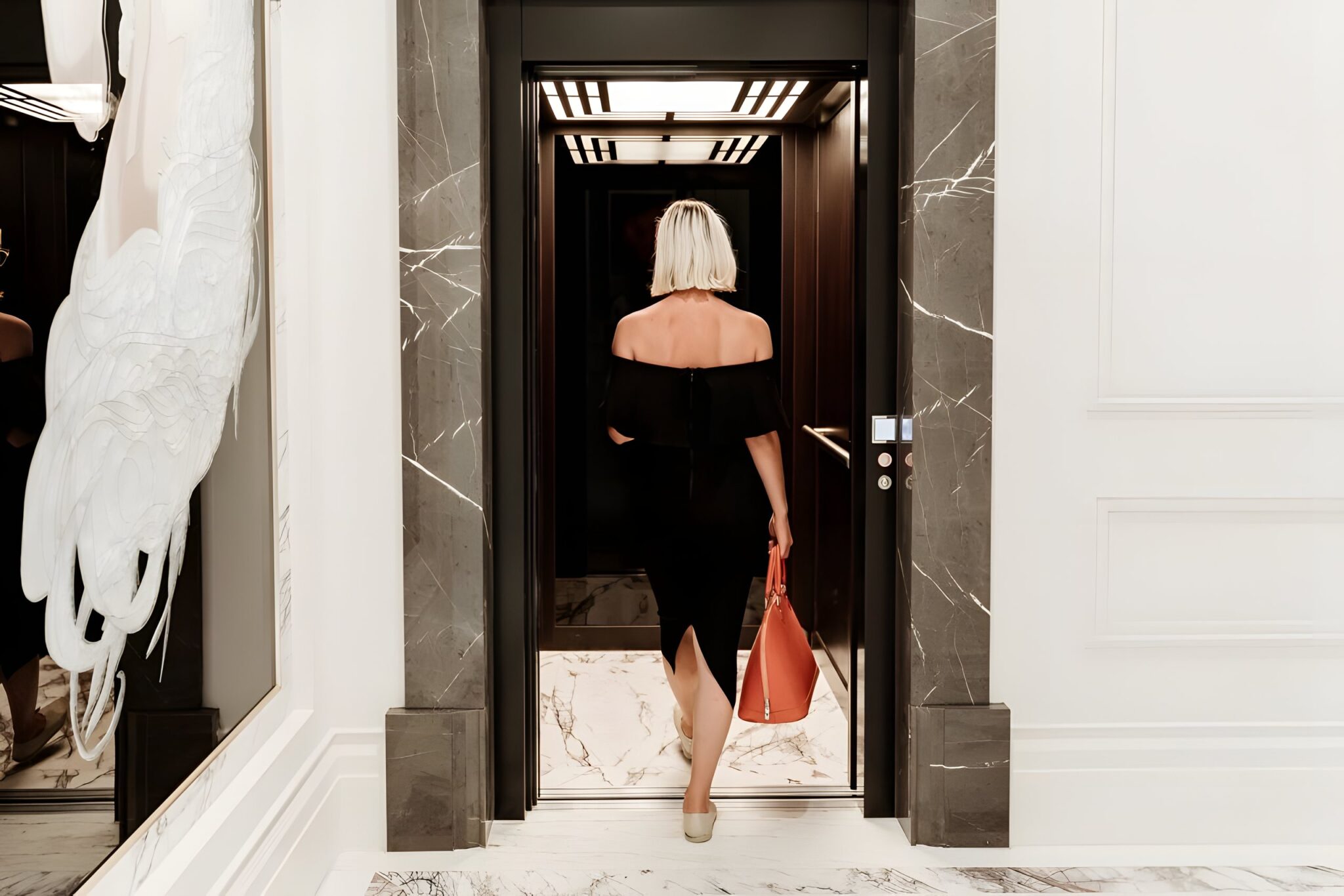 Woman in a black dress walking into a lift framed by a grey marble surround in a white panelled hallway, Leura