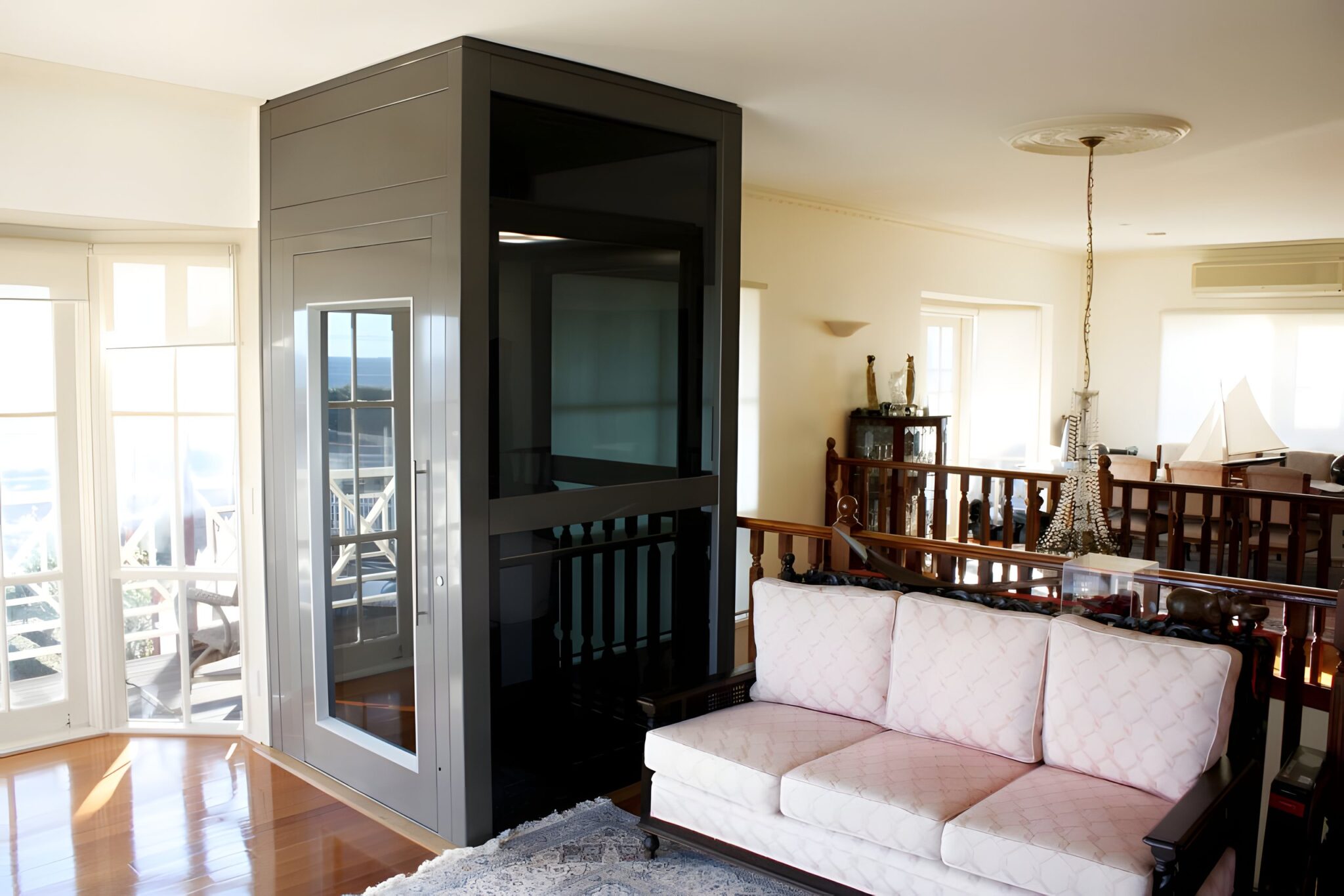 Grey lift shaft with a glass-fronted door beside a sofa and timber balustrade in a Hampton living room