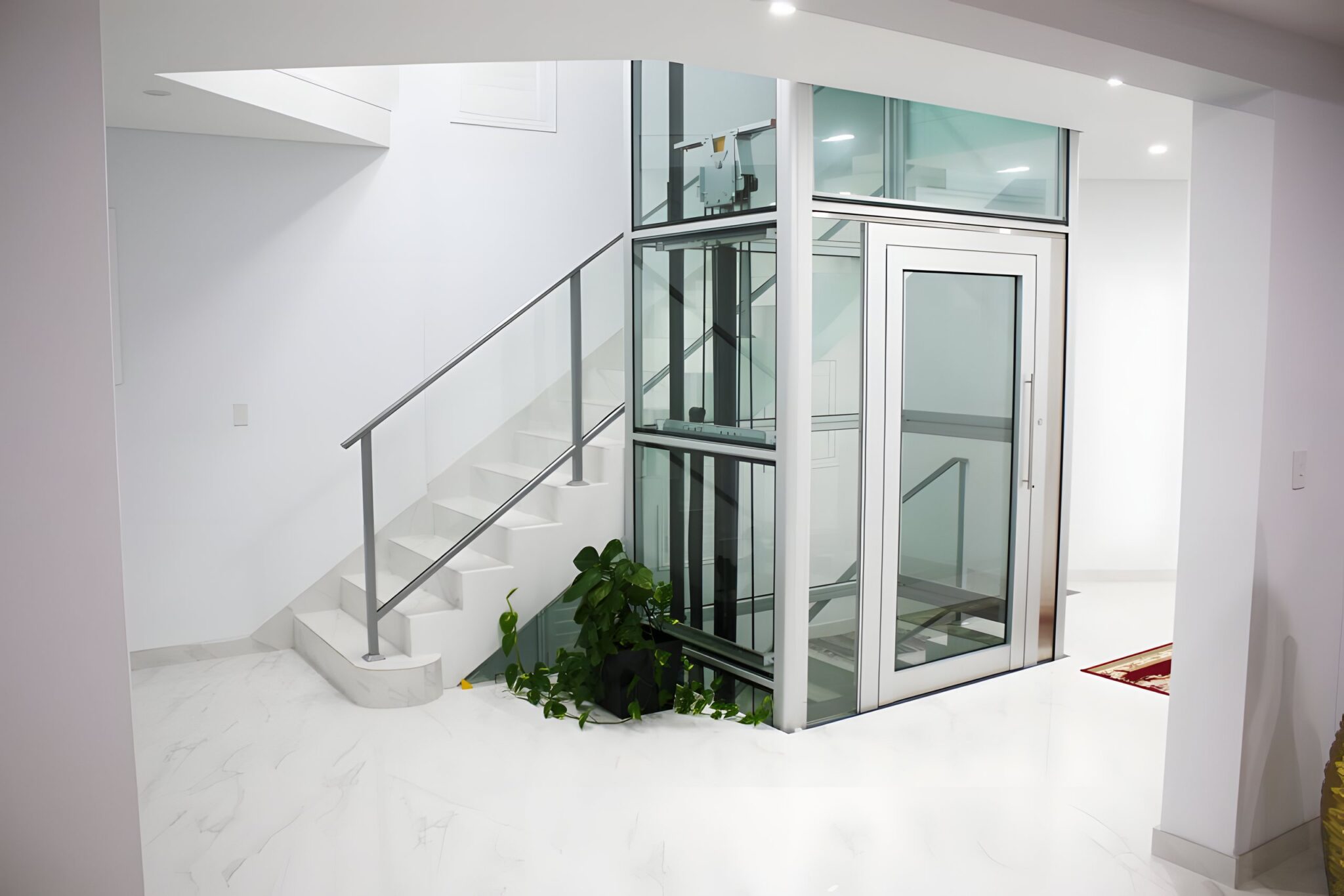 Glass-shaft home lift beside a white marble staircase with a glass balustrade in a Los Angeles home