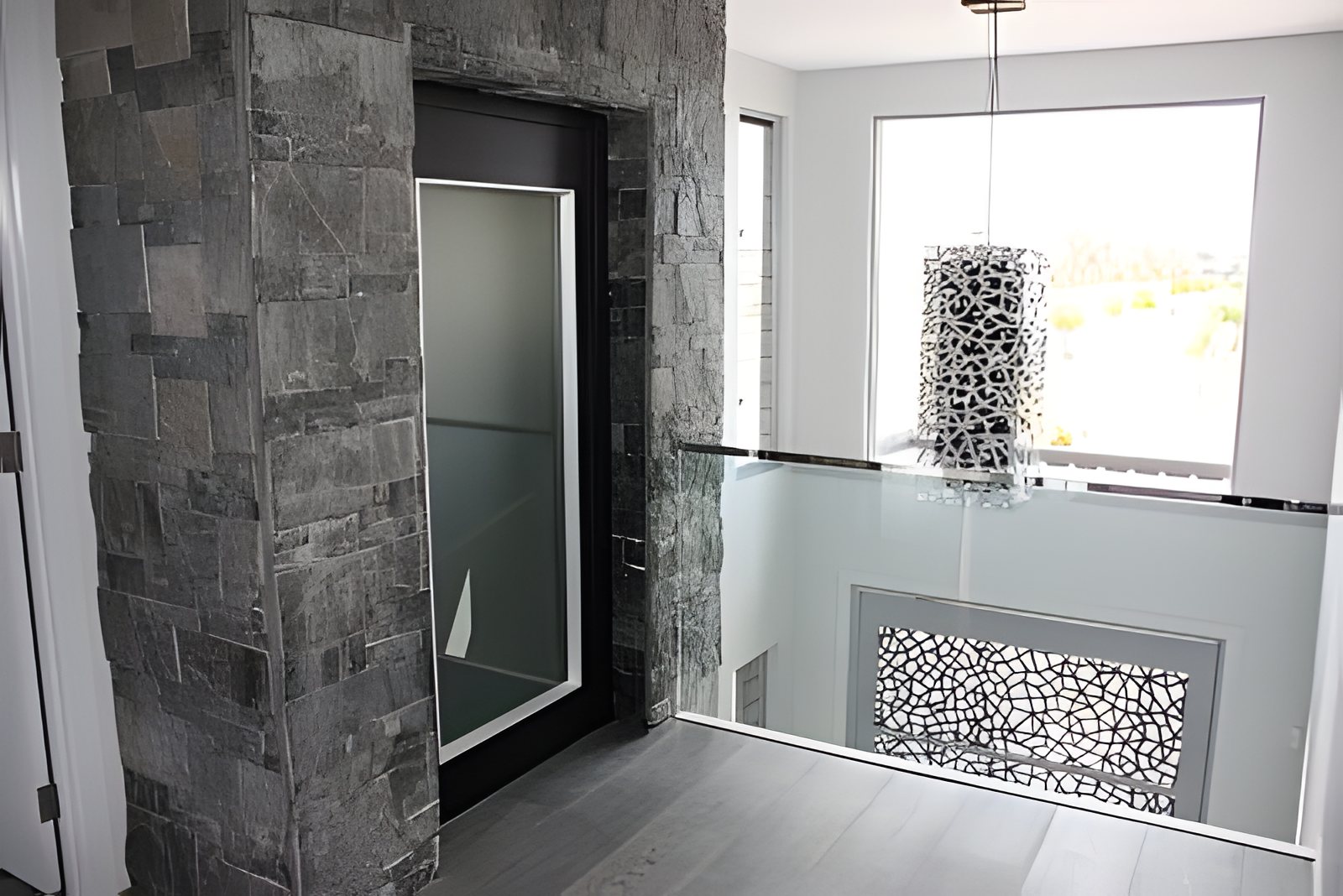 Landing with a stone clad lift surround, black framed frosted glass lift door and a pendant light in a Gold Coast home