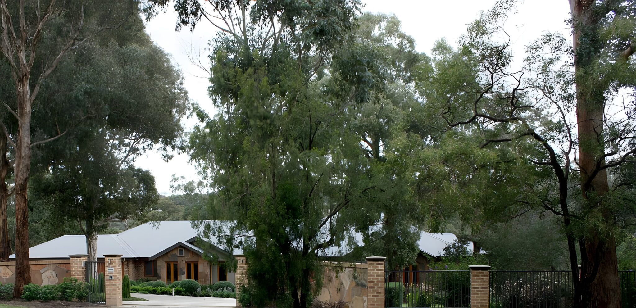 House with a pitched grey roof behind tall gum trees and a brick and stone front fence in Plenty