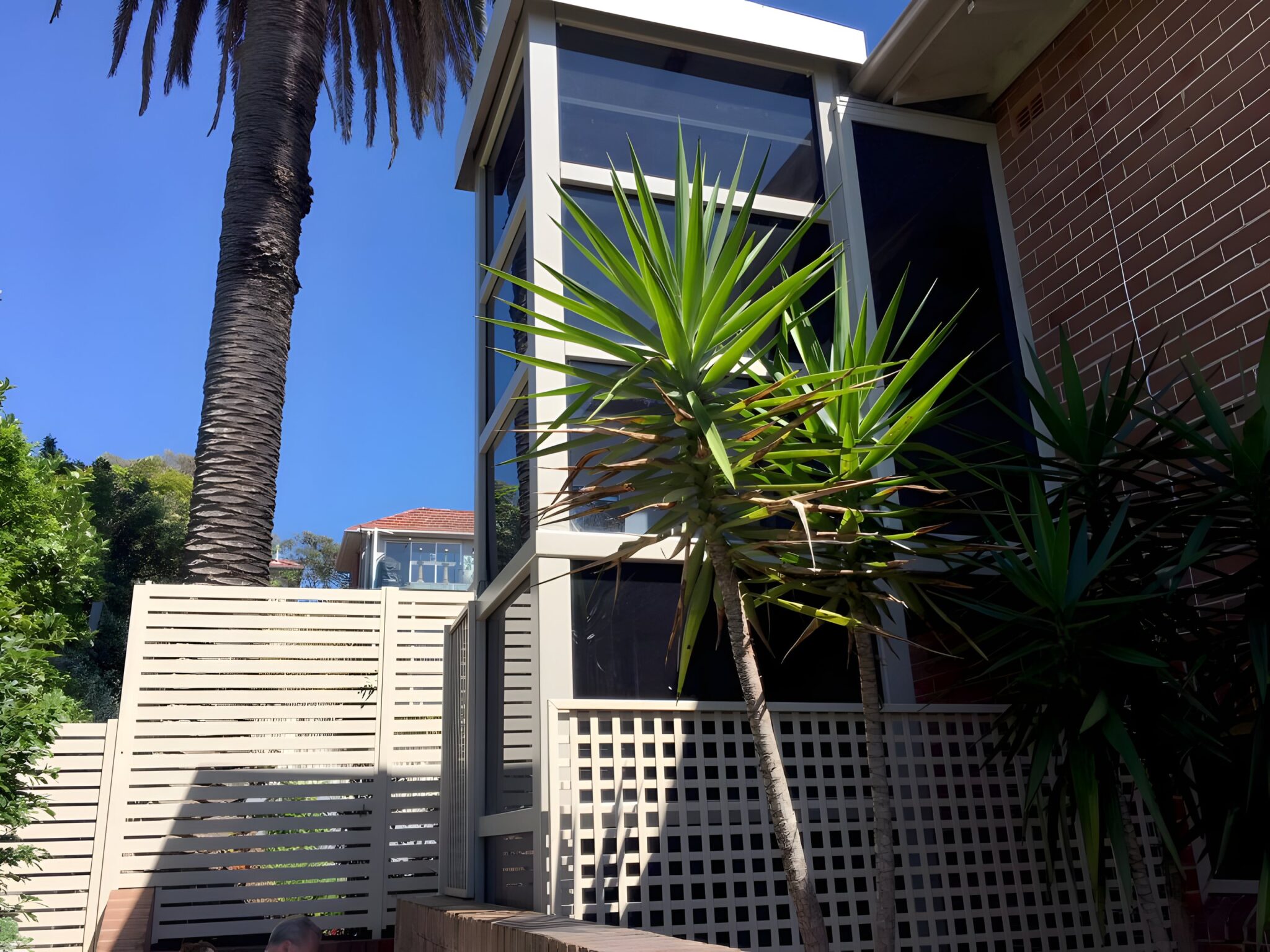 Glass lift shaft rising beside a brick wall and palm trees in Mosman