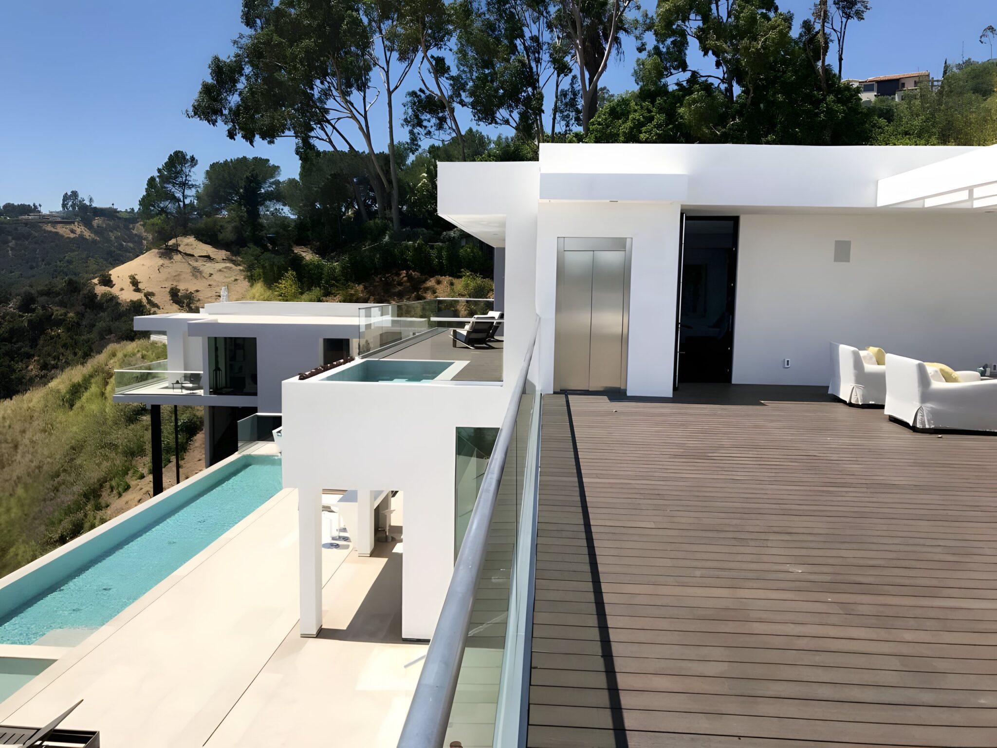 Brushed stainless steel lift door on a white Bel Air estate, beside a timber deck, glass balustrade and infinity pool above the hills