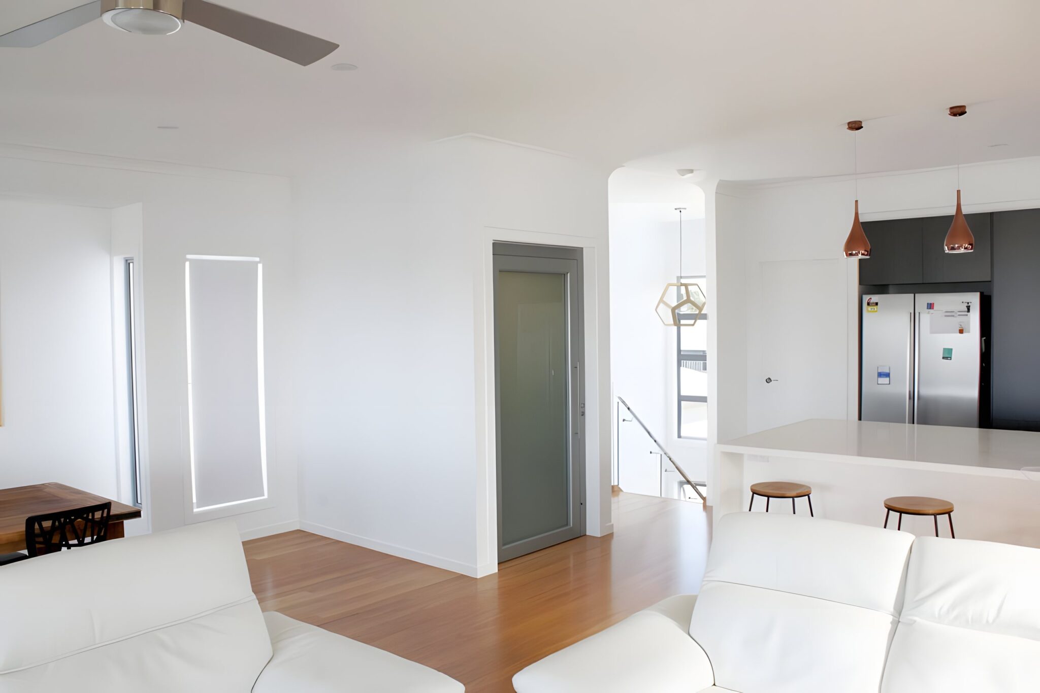 Open-plan living area in an Ormiston home with a grey-framed milk-white glass lift door opening onto the timber floor, white sofas and a kitchen with copper pendant lights