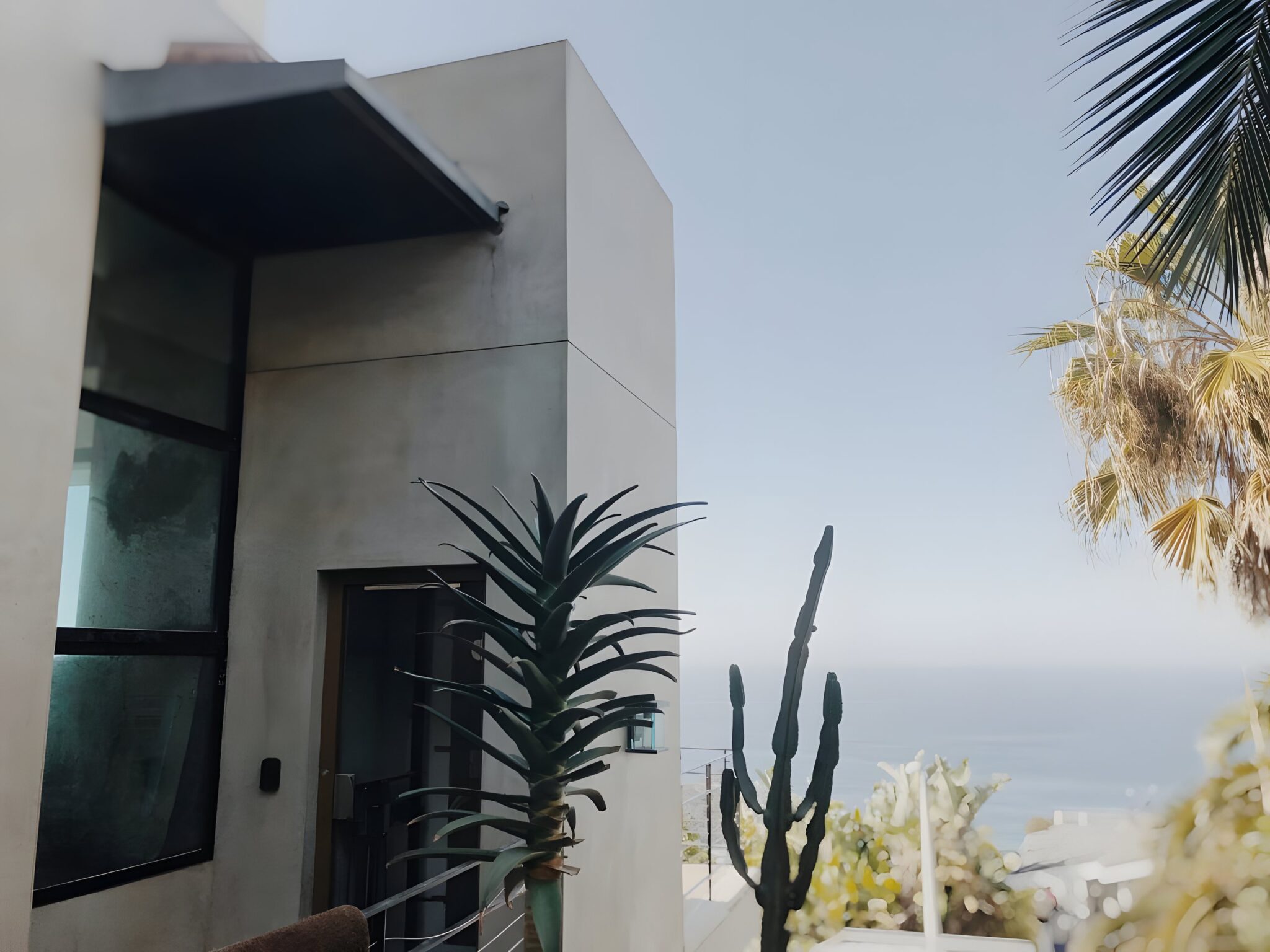 Concrete rendered lift tower with a dark glass door among palms and cacti, with the ocean beyond