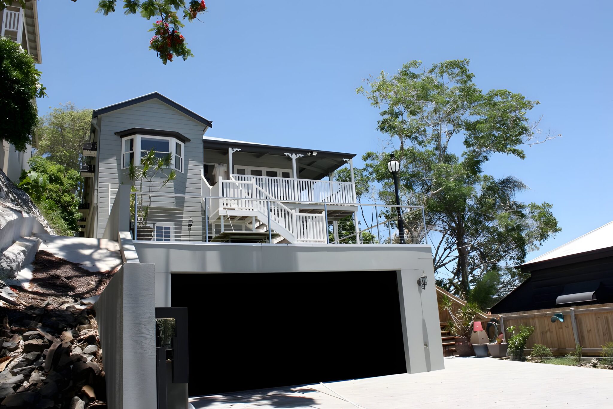 Grey two storey Queenslander home on a hillside above a double garage, with a white verandah and balustrade, under a blue sky