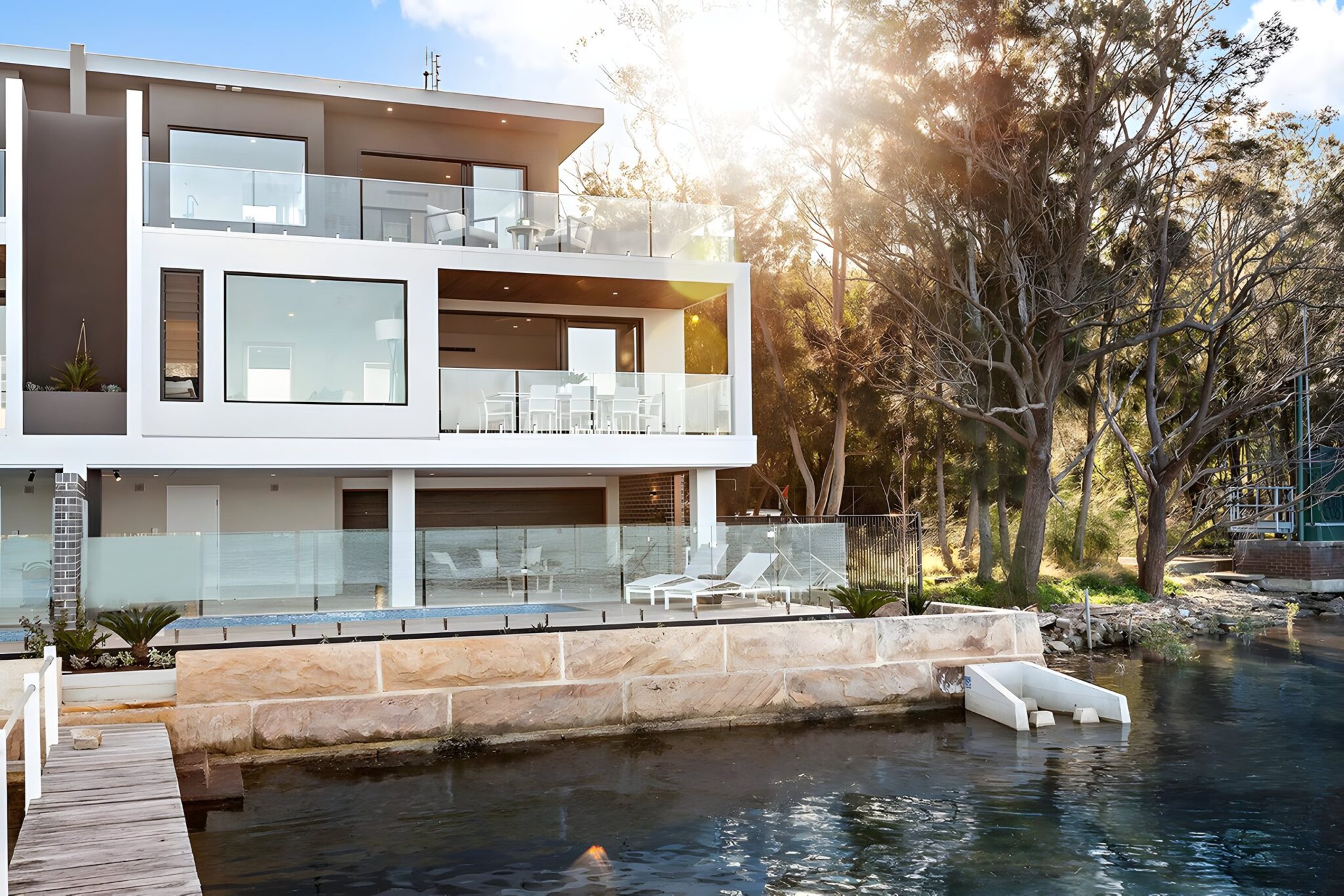 Three-storey white waterfront home with glass balconies and trees beside the water