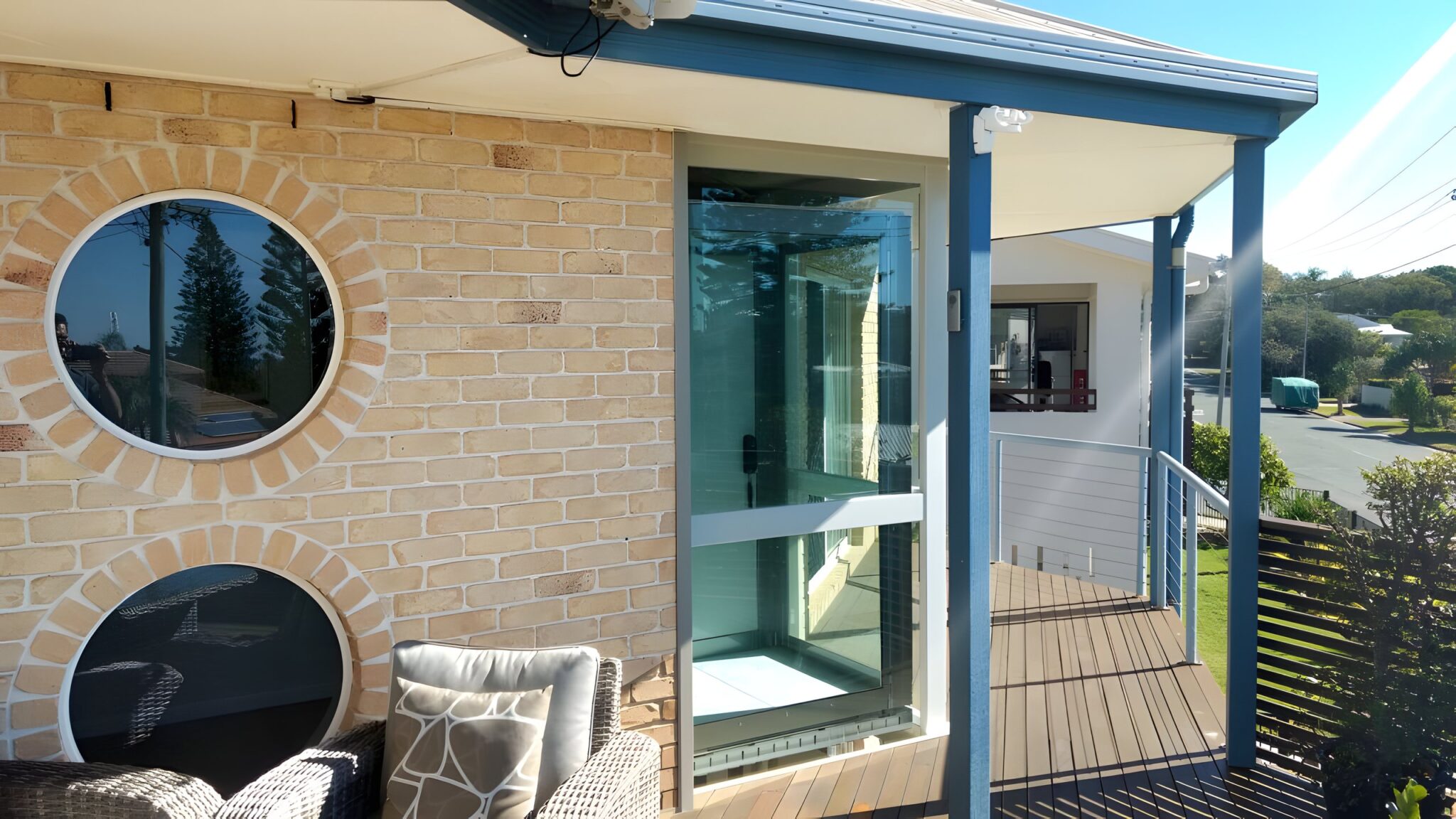 Glass lift shaft with a blue frame beside round windows on a brick wall, under the verandah of a Sunshine Coast home