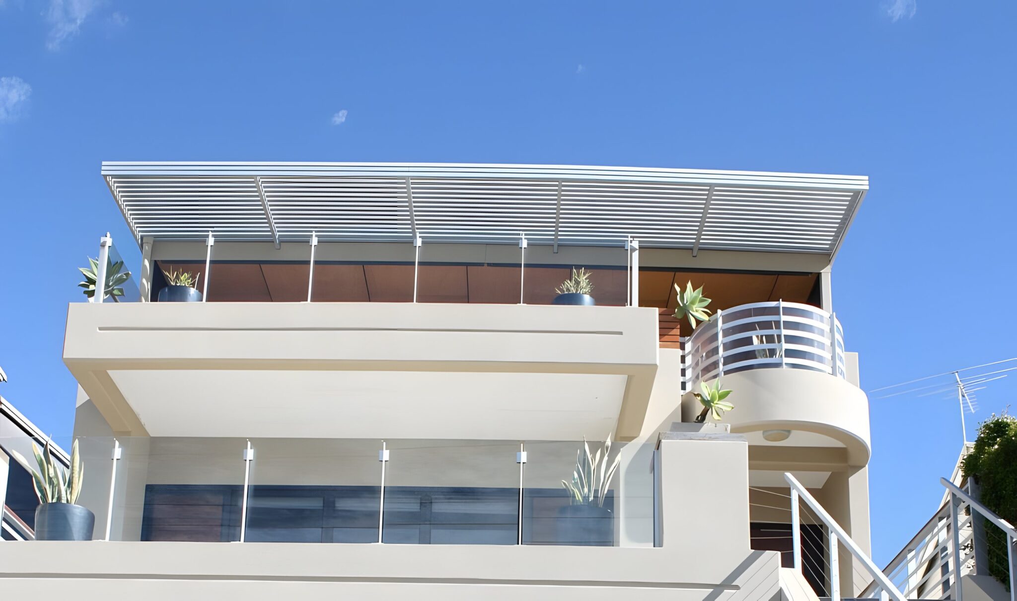 Multi level Bulimba home with a louvred roof, glass balustrades and a curved balcony against a blue sky
