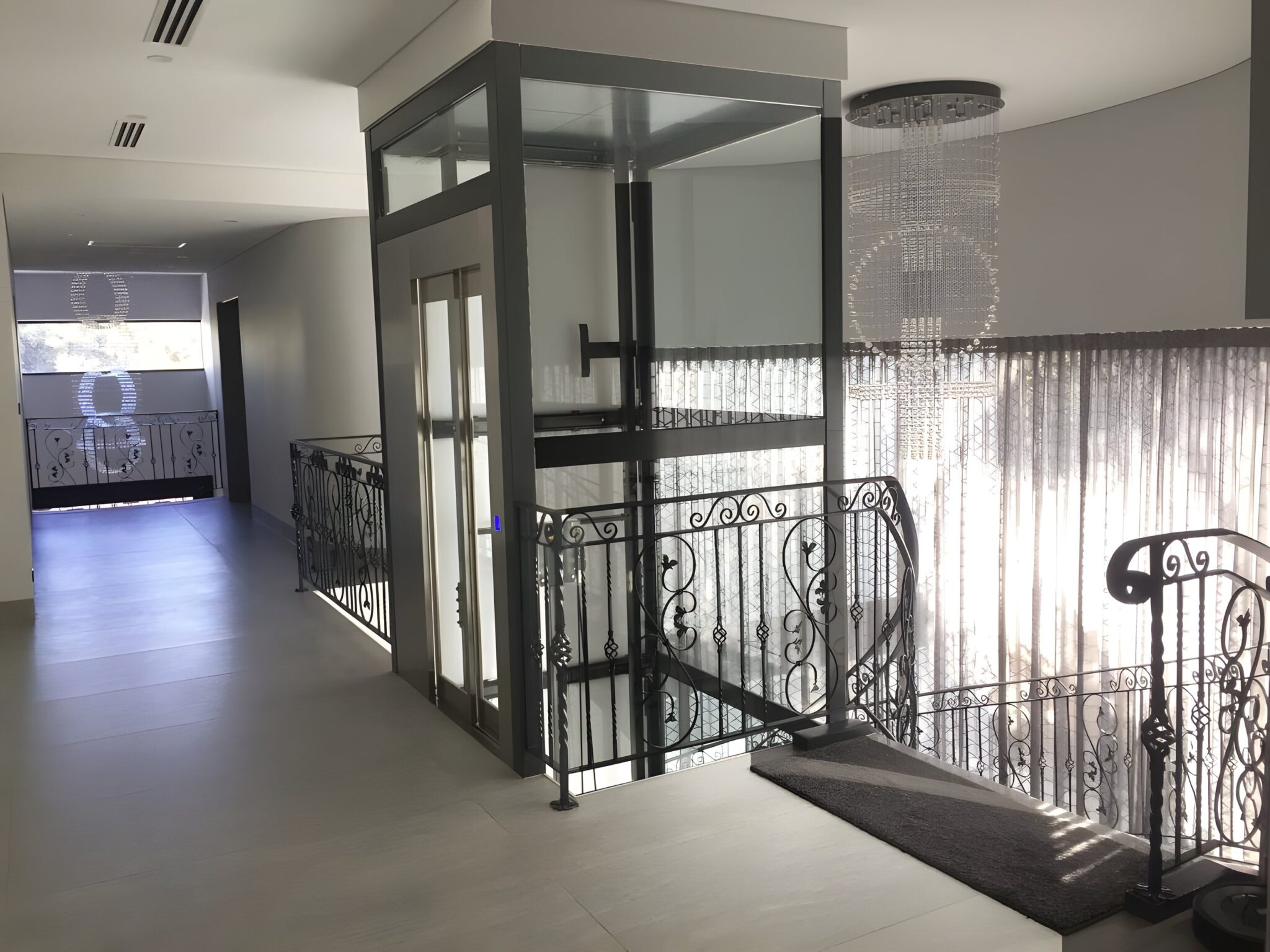 Glass home lift beside a curved wrought iron balustrade on an upper landing with a hanging crystal chandelier
