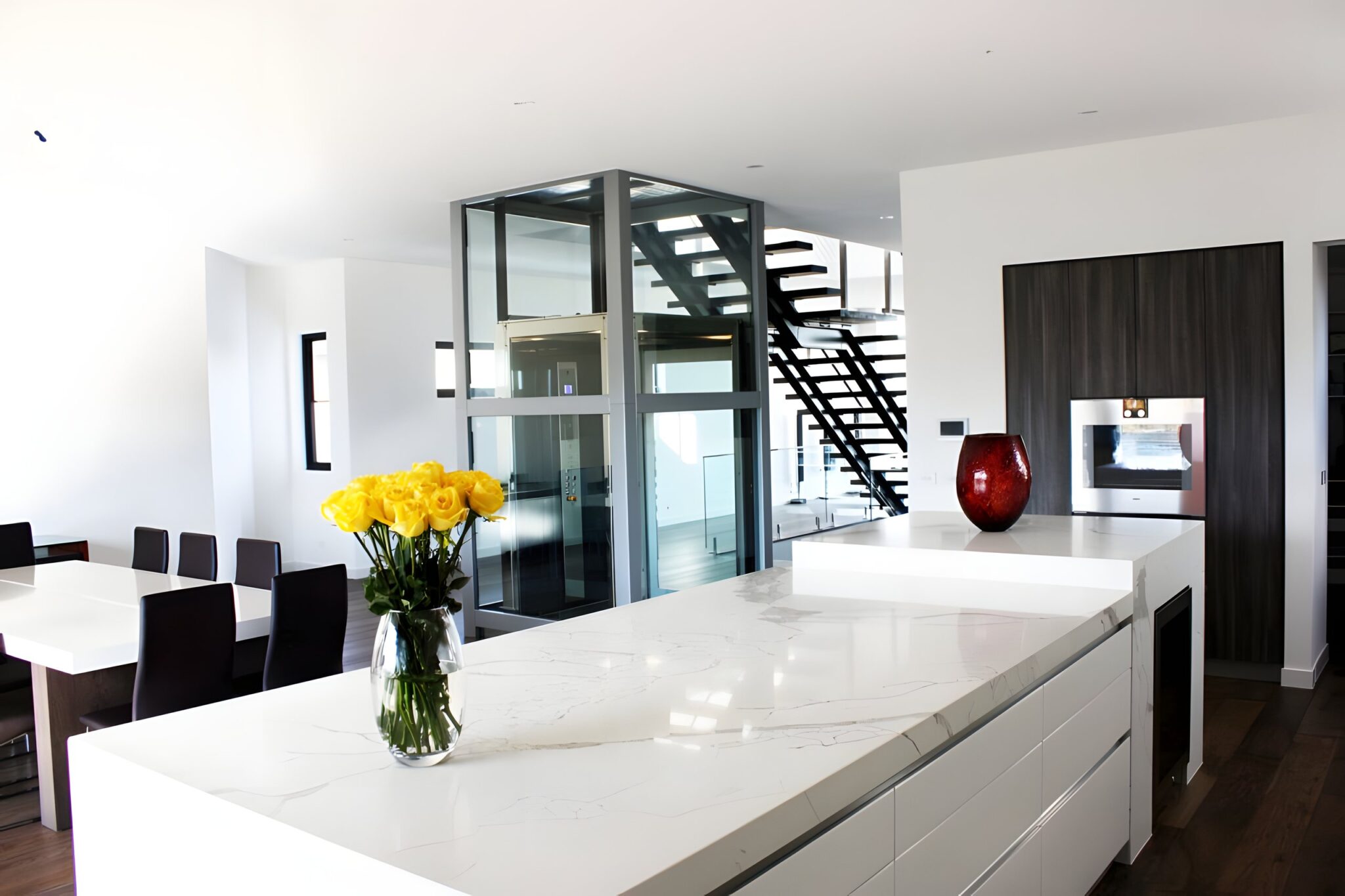 Glass-shaft lift with a polished stainless steel and glass cabin standing in an open-plan kitchen and dining area, with a black stair behind and yellow roses on a marble bench in the foreground