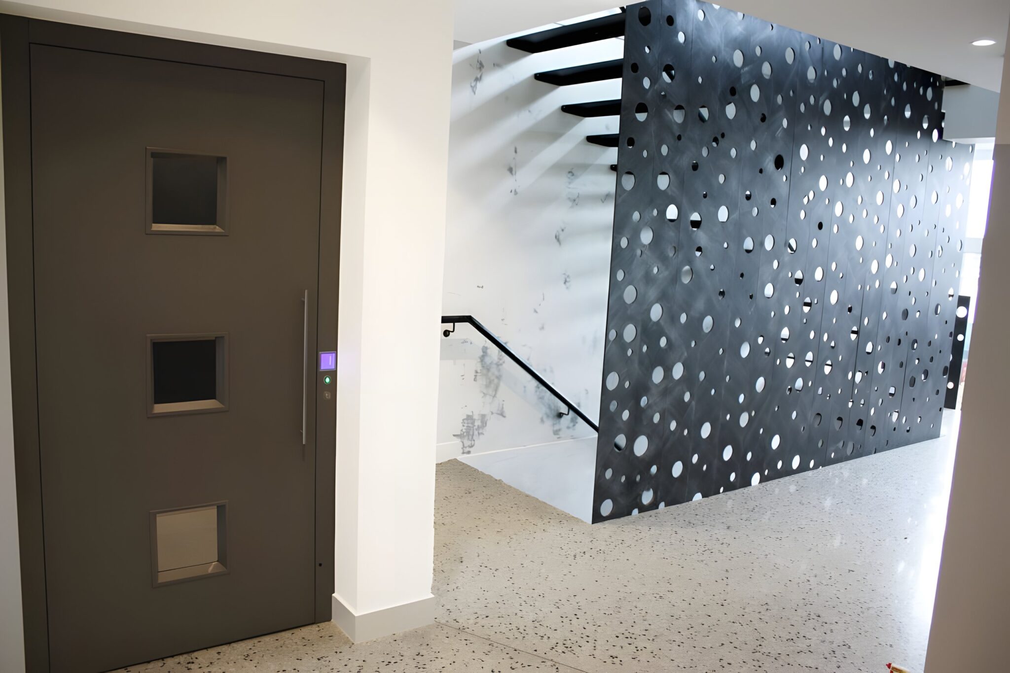 Dark grey lift door with three small windows beside a perforated black steel screen in Maribyrnong
