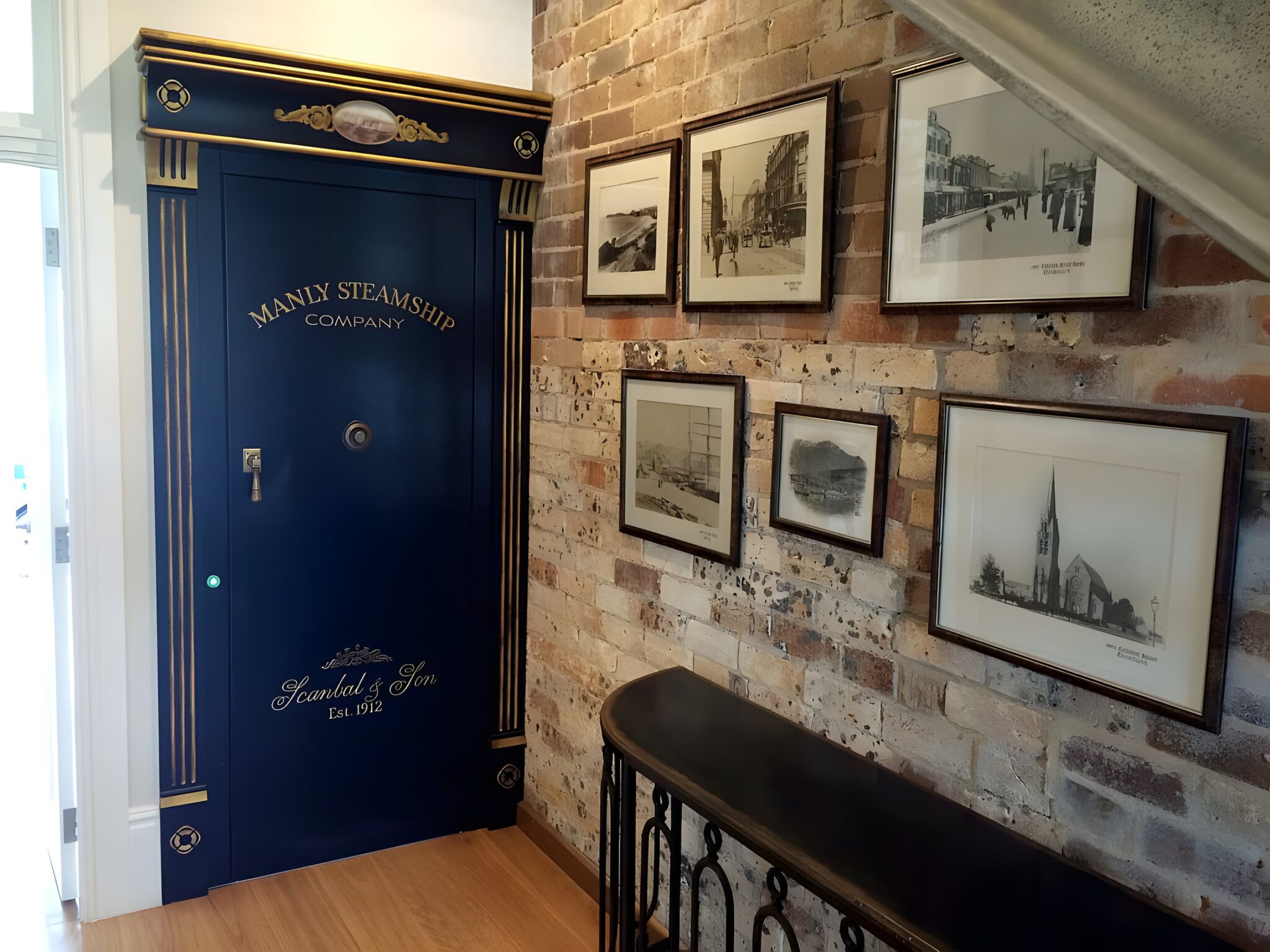 Navy vault-style lift door lettered Manly Steamship Company beside an exposed brick wall hung with framed photos