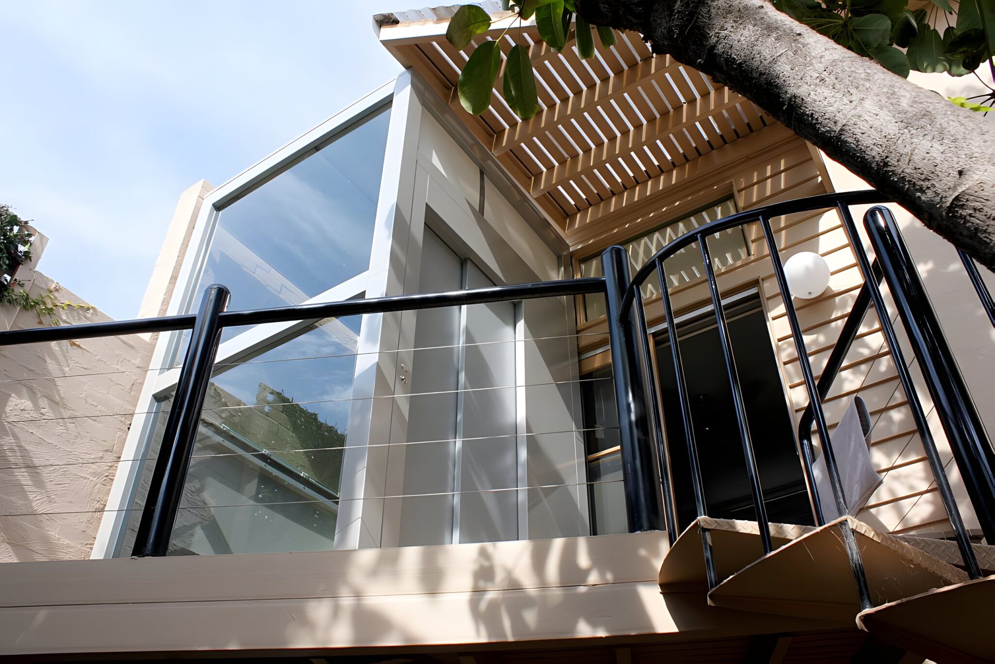 Glass-shafted E2 lift seen from below beside a timber pergola and black balustrade in a Melbourne courtyard