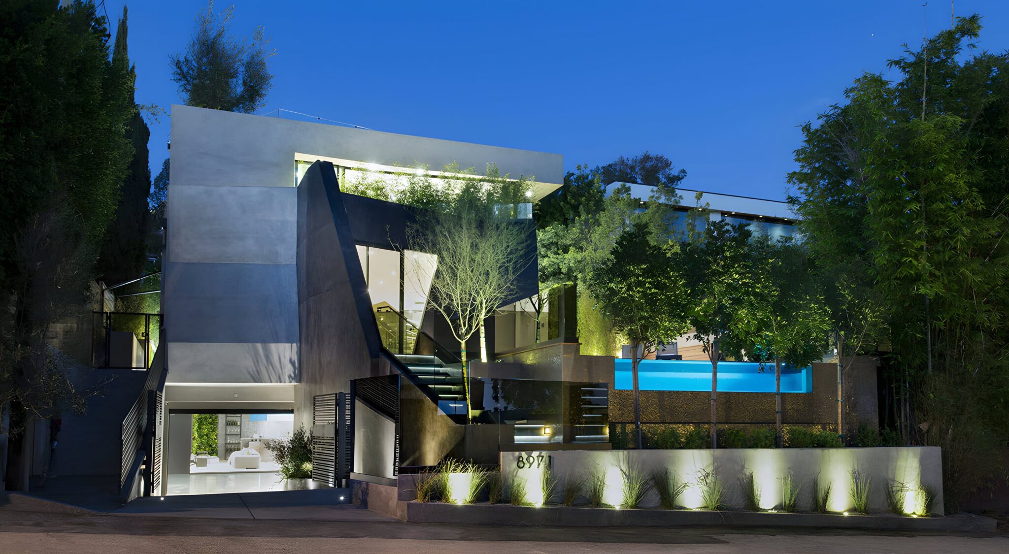 Modern angular home with a lit pool above the garage at dusk, surrounded by trees, the Sunset Strip home with the four-floor E1 lift