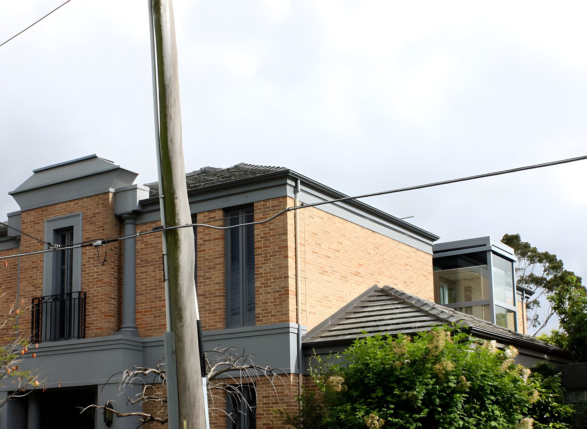 Brick two-storey Toorak home with a grey upper level and a glass lift shaft on the roofline, seen from the street
