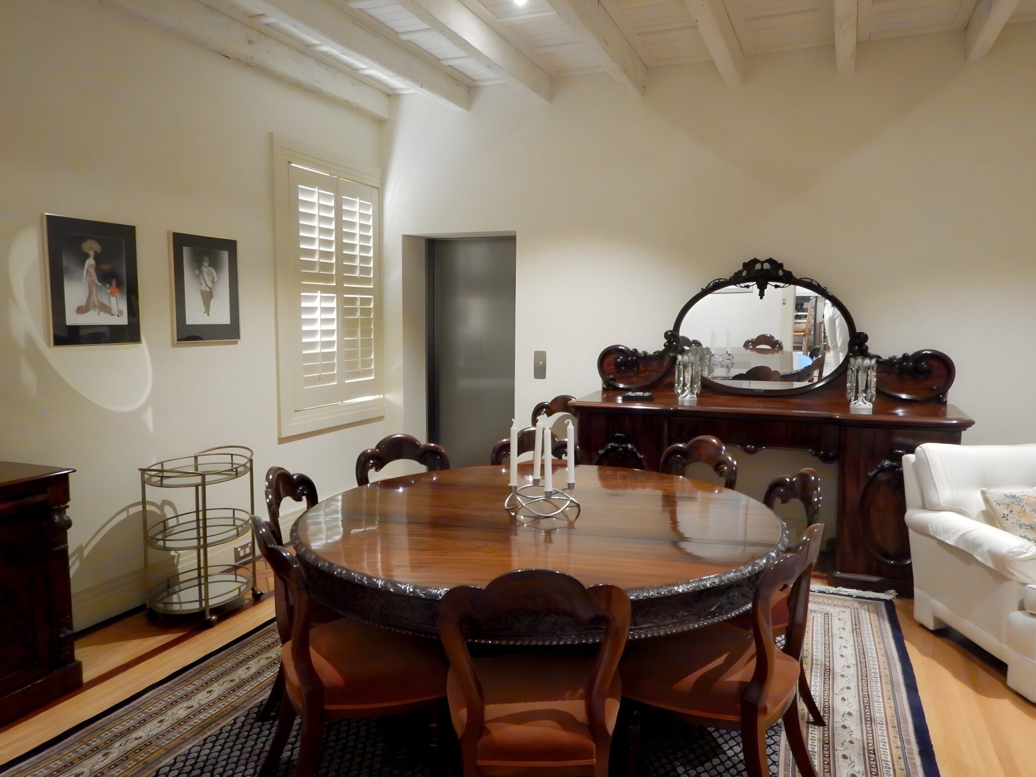 Dining room with a round timber table and antique sideboard, with the lift door set into the wall beside the window shutters